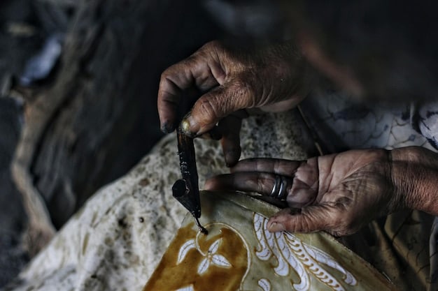 A close-up of a volunteer carefully cleaning dust from an old, intricately carved wooden statue in a dimly lit museum storage area. The volunteer wears gloves, and the statue's details are highlighted by soft artificial light.
