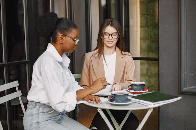 Two professionals are engaged in a lively conversation over coffee, with open body language and genuine smiles, in a bright, modern office cafe setting.